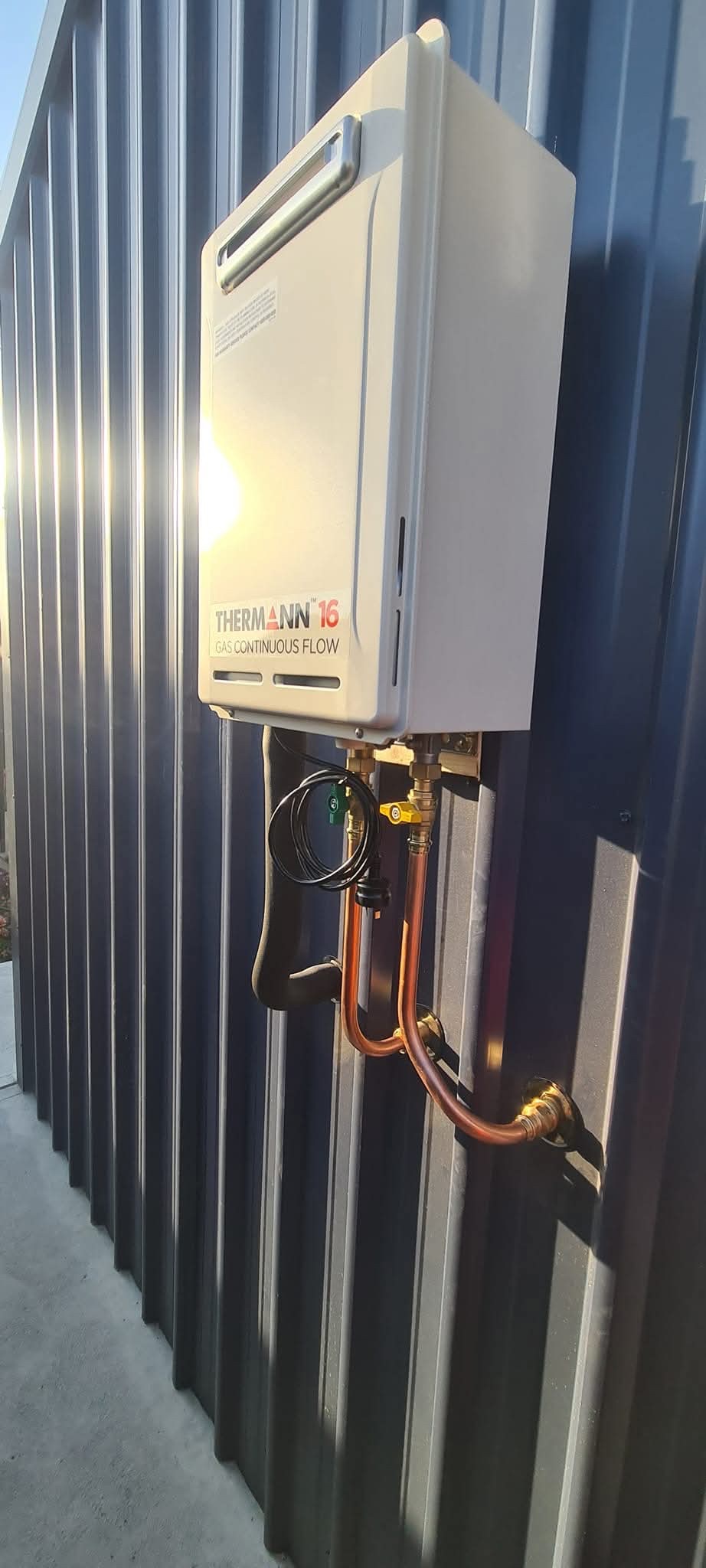 Completed Thermann gas continuous-flow hot-water installation on a dark exterior wall with visible copper pipework